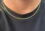 A close-up of a person's neck wearing a delicate, thin chain necklace with small, multi-colored round beads in rainbow shades, paired with a black crewneck shirt.