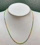 A delicate beaded chain necklace with small multicolored beads in yellow, green, blue, and pink, displayed on a white jewelry bust against a textured dark green background.